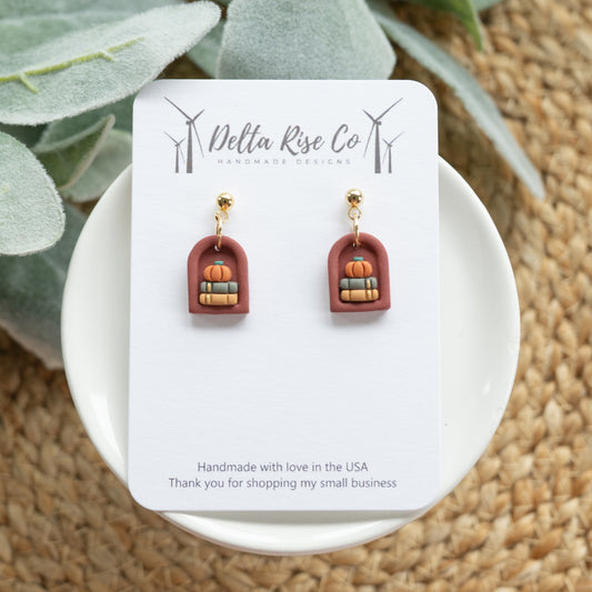 Handmade clay mini autumn bookshelf earring featuring a stack of books topped with a small pumpkin, perfect for fall, autumn, or Halloween style. This cozy seasonal accessory combines book lover charm with festive autumn details, making it a unique piece of polymer clay jewelry. Ideal for readers, teachers, or anyone who loves autumn accessories, fall fashion, and Halloween-inspired handmade jewelry.